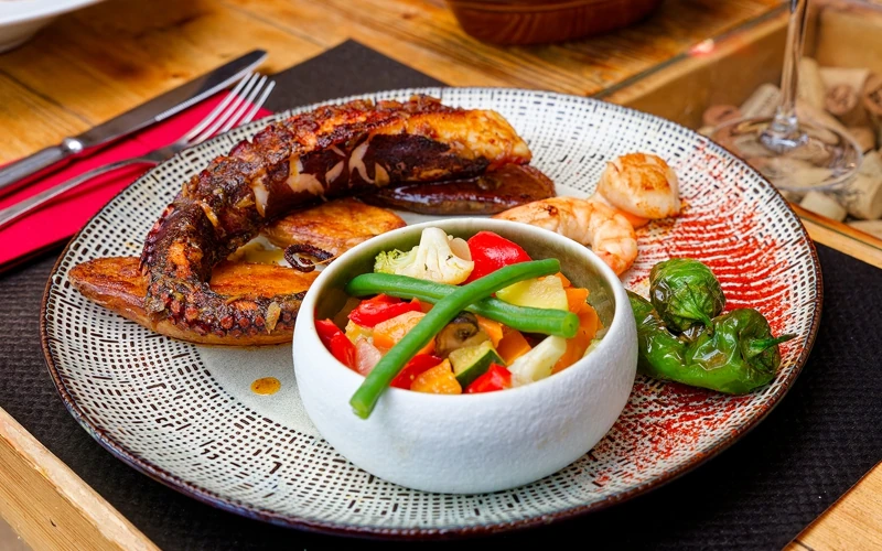 Appétissante assiette dans laquelle sont dressés des morceaux de poulpe à la Pericana avec ses légumes croquants, crevettes et pimientos de padron